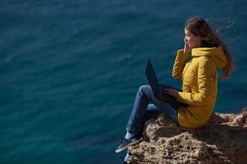 A freelancer girl is working typing on a laptop and talking on a mobile phone with a beautiful view of the open air sea sky. Traveling with a computer. Online dream job.