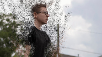 A young Caucasian man wearing a black t-shirt and glasses stares straight ahead from a side angle as his body disperses into a bunch of tiny particles floating around him sucked to the sky above him.