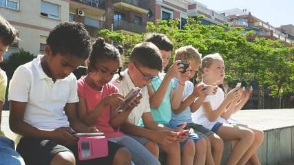 Wall Mural - Group of positive kids sitting at urban street with mobile devices 