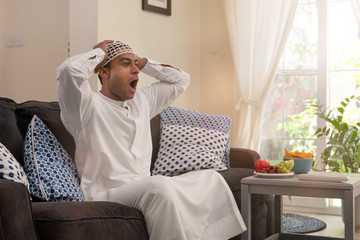 Wall Mural - Excited anxious Middle Eastern, Emarati, Saudi, Moroccan man wearing a traditional kandura and prayer cap watching TV on his sofa at home