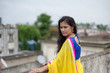 © abir - Young and beautiful Indian Bengali brunette woman in Indian traditional dress yellow sari and blue blouse is standing thoughtfully leaning on rooftop wall under blue sky with clouds. Indian lifestyle