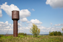 Old Water Tower In The Countryside. The Metal Tower, Covered With Rust, Reinforced With Metal Cables, Stands In The Middle Of A Green Meadow On A Sunny Day Against A Blue Sky With Clouds.
