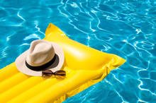 Sun Hat And Sunglasses Resting On Bright Yellow Inflatable Raft Floating In Blue Swimming Pool