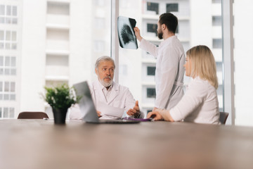 Young doctor stands by the window with an X-ray. Two colleagues of the doctors sit at the table and watch the young doctor. Concept of team medical work.