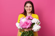 © sementsova321 - Indoor shot of nice young woman holding beautiful fresh blossoming bouquet of white and pink peony flowers, lady wearing yellow dress, posing isolated over rose background, looking smiling at camera.