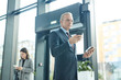 © AnnaStills - Horizontal low angle portrait of mature Caucasian businessman standing in airport holding his purse and boarding pass watching something on smartphone