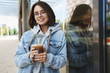 Young attractive girl in glasses, denim jacket, having casual walk in city, enjoying weekends, drinking ice latte, leaning on building wall and smiling camera with happy relaxed expression