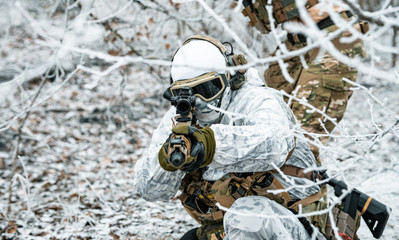 Airsoft man in white camouflage uniform aims at the sight. Soldier in the winter forest between branches. Horizontal photo.