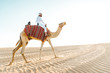 © oneinchpunch - Man wearing traditional clothes, taking a camel out on the desert sand, in Dubai