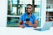 © Carlos David - Portrait of a friendly male doctor or nurse wearing blue scrubs uniform and stethoscope sitting at desk with laptop in hospital checking mobile phone