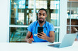 © Carlos David - Portrait of a concerned male doctor or nurse wearing blue scrubs uniform and stethoscope sitting at desk with laptop in hospital checking mobile phone