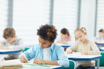 Horizontal medium close up shot of concentrated African American boy sitting at desk in classroom writing composition, copy space