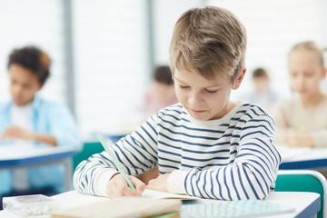 Caucasian boy wearing striped sweatshirt sittng at desk in classroom doing lesson task in his notebook, horizontal medium close up portrait, copy space