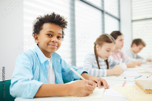 Portrait of cheerful African American boy sitting at school desk with his classmates looking at camera smiling, copy space