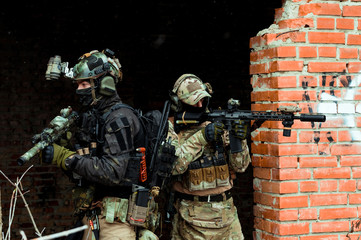 Two men in camouflage cloth and black uniform with machineguns stand beside brick wall. Soldiers with muchinegun aims aiming from around the corner