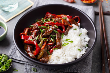 Wall Mural - Beef and vegetables stir fry with white rice in a black bowl. Grey background. Close up.