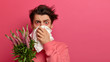 © wayhome.studio - Seasonal allergy and health problems concept. Unhealthy man blows nose in tissue, being sensitive to allergens, suffers from bad symptoms, holds blossom plant, covers face with handkerchief.