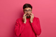 Scared horrified man stares at camera, has phobia, feels afraid to death, has alarmed face expression, wears glasses, grabs face, isolated on pink background. Frightened male hears wild news