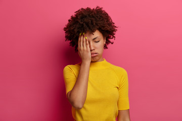 Wall Mural - Exhausted dark skinned young woman covers half of face, sighs from tiredness, has sleepy expression, closes eyes, wears yellow t shirt, poses over pink background. Female feels bored and tired