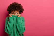 Positive Curly Young Woman Pulls Green Sweater Over Head, Has Fun And Hides Face, Looks From Underneath, Disappears In Her Clothes, Gazes Gladfully, Isolated On Pink Background, Copy Space Area