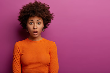 Indoor shot of shocked curly haired woman horrified to find out about awful accident, dressed in orange jumper, stands against purple background, surprised by sudden news. Human reaction concept