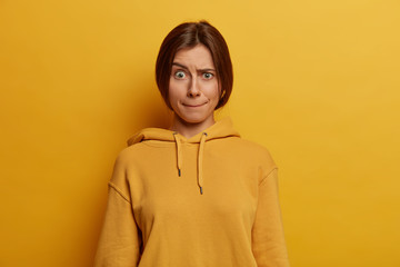 Poster - Beautiful dark haired young European woman presses lips, raises eyebrows, has worried face expression, wears casual hoodie, poses against yellow background, has embarrassed look. Monochrome shot.