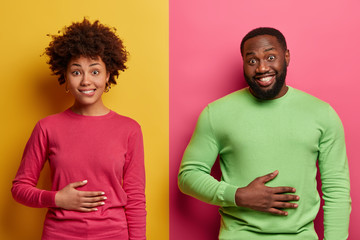 Wall Mural - Pleased ethnic young woman and man keep hands on stomach, feel satiety after eating tasty nutritious dinner, smile positively, happy not to be hungry, pose against yellow and pink background