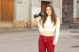 Fototapeta Sypialnia - Portrait of attractive caucasian young woman model holding hair dryer, in the city, white top and red pants. Place for your text in copy space.