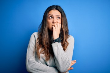 Young beautiful brunette woman wearing casual sweater standing over blue background looking stressed and nervous with hands on mouth biting nails. Anxiety problem.