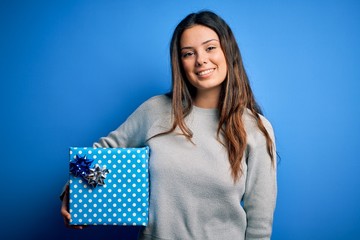 Young beautiful brunette woman holding birthday gift over isolated blue background with a happy face standing and smiling with a confident smile showing teeth