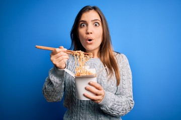 Sticker - Young woman eating asian noodles from take away box using chopstick over blue background scared in shock with a surprise face, afraid and excited with fear expression