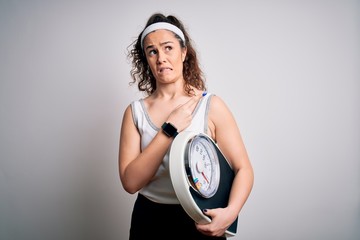 Sticker - Young beautiful woman with curly hair holding weighing machine over white background Pointing aside worried and nervous with forefinger, concerned and surprised expression