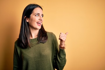 Wall Mural - Young brunette woman with blue eyes wearing green casual sweater over yellow background smiling with happy face looking and pointing to the side with thumb up.