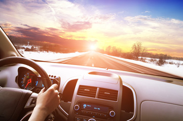 Canvas Print - Car dashboard with driver's hand on the steering wheel against a winter road in motion and a sky with a sunset