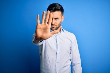 Young handsome man wearing elegant shirt standing over isolated blue background doing stop sing with palm of the hand. Warning expression with negative and serious gesture on the face.