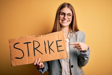 Young beautiful redhead woman holding banner with strike message over yellow background very happy pointing with hand and finger