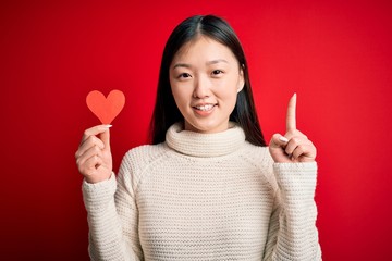 Young asian woman holding romantic red heart paper shape over red isolated background surprised with an idea or question pointing finger with happy face, number one