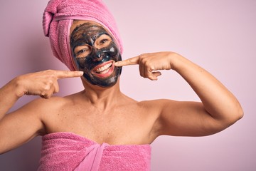 Wall Mural - Middle age brunette woman wearing beauty black face mask over isolated pink background smiling cheerful showing and pointing with fingers teeth and mouth. Dental health concept.