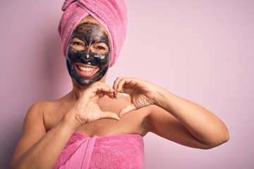 Wall Mural - Middle age brunette woman wearing beauty black face mask over isolated pink background smiling in love doing heart symbol shape with hands. Romantic concept.