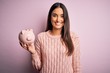 Young beautiful brunette woman holding piggy bank saving money for retirement with a happy face standing and smiling with a confident smile showing teeth