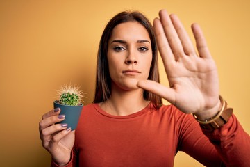 Young beautiful brunette woman holding small cactus pot over yellow background with open hand doing stop sign with serious and confident expression, defense gesture