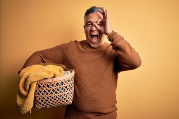 Poster - Middle age handsome grey-haired man doing housework holding wicker basket with clothes with happy face smiling doing ok sign with hand on eye looking through fingers