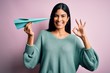 © Krakenimages.com - Young beautiful hispanic woman holding paper plane and communication and travel message doing ok sign with fingers, excellent symbol