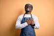 Young african american cooker man wearing apron and over isolated yellow background smiling with hands on chest with closed eyes and grateful gesture on face. Health concept.