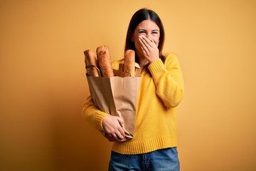 Poster - Young beautiful woman holding a bag of fresh healthy bread over yellow background laughing and embarrassed giggle covering mouth with hands, gossip and scandal concept