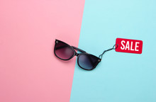 Fashionable Sunglasses With Red Sale Tag On Pink Blue Pastel Background. Top View. Discount. Minimalism