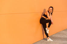 Attractive Sexy Girl With Fit Body In Tight Sportswear, Black Pants And Top, Leaning Against Orange Wall And Smiling To Camera, Living Healthy Sport Life, Full Of Energy. Isolated On Advertising Area
