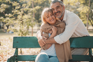 Wall Mural - Relaxing,family,free time,age,tourism,travel and people concept.Senior couple hugging and Happy mature people in winter clothes embracing on bench.Elderly couple sitting on bench in autumn park.