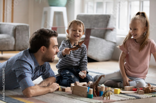 Fotomural Happy little boy playing with toy dinosaur, having fun with smiling father and elder cute sister in living room