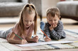 © fizkes - Playful adorable blonde girl lying on carpet floor with younger brother, drawing pictures together in paper album. Interested two little children siblings involved in creative activity hobby at home.
